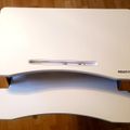 A white laptop stand with two curved tiers on a wooden floor, branded "MOUNT-IT" with a slot for holding a device on the upper level