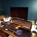 A cluttered wooden desk setup with a large monitor, gaming laptop with RGB keyboard, VR headset, and various tech accessories in a room with teal walls