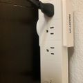 White power strip with multiple outlets and USB ports mounted on wall with black electrical cord plugged in