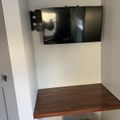 A flat-screen TV mounted on an articulating wall bracket above a wooden shelf, with a patterned cushioned stool below in a white-walled room