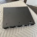 A black rectangular electronic device or component with mounting holes and connectors along one edge, placed on a woven placemat on a wooden table with a chair visible in the background