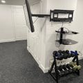 A basement room with white walls and gray carpet featuring a wall-mounted TV, floating shelves with electronics, and a dumbbell weight rack in the corner
