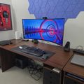 A modern computer workstation with a large curved monitor displaying a colorful tunnel-like screensaver, wooden desk with keyboard and mouse, gaming controller, speakers, and blue hexagonal acoustic panels on the wall above