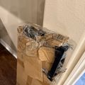 A cardboard box wrapped in plastic sits on a wooden floor against white walls and baseboard trim. The box appears to be a delivery package with a barcode label visible, and contains what looks like a black item partially wrapped in plastic inside.