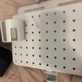 White perforated plastic basket or container with handles and mounting hardware, placed on a beige fabric surface