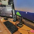A wooden desk setup with a dual monitor gaming configuration, including a left monitor displaying a game interface, a right larger monitor showing a mountain landscape, a black keyboard, gaming mouse with stand, speakers, and various desk accessories including a yellow cup and pink scissors.