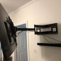 A flat screen TV mounted on a black articulating wall mount bracket that extends from the wall, with cables visible and a media device shelf below