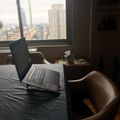 A laptop computer sits open on a bed next to a brown leather chair, with a large window showing a city skyline view of tall buildings in the background.