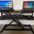A height-adjustable standing desk with two laptops open on the upper surface and a keyboard on the lower shelf, set up in an office environment with carpeted flooring