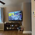 Living room with large TV mounted on wall above dark wooden entertainment center, showing what appears to be a sports broadcast with celebrating people, hardwood floors, and wicker basket with stuffed animals nearby