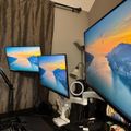 A home office setup with three computer monitors displaying a scenic mountain and water landscape, along with a desk lamp, microphone, and other office equipment on a dark desk near a window with curtains.