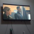 A flat screen TV mounted on a wall displaying a movie or TV show with two people in what appears to be a dramatic scene, with entertainment equipment visible on a shelf below