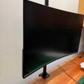 A curved black computer monitor mounted on a wooden desk with a black stand, displaying a dark screen with a green gradient at the bottom. A keyboard is partially visible in the foreground on the right side.