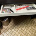 Open desk drawer containing office supplies including a magnifying glass, calculator, red pen, and small decorative items, with patterned tile flooring visible below