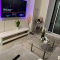 A modern living room with a wall-mounted TV displaying a streaming service, white floating TV stand with potted plant, glass coffee table on a geometric patterned rug, decorative shelf with cosmetics and plants, and a sliding glass door leading to a balcony. Purple ambient lighting illuminates the wall behind the TV.