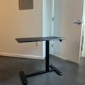 A height-adjustable standing desk with black tabletop and metal base positioned in a room with white walls and gray tile flooring