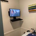 A wall-mounted computer monitor and keyboard setup in a corner of a room with white walls and decorative molding, with a colorful striped artwork visible on the wall.