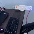 A desk setup with a black laptop, wireless keyboard, mouse, and white power strip with USB ports mounted above the desk surface
