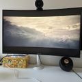 Computer monitor displaying a cloudy sky scene with webcam mounted on top, keyboard below, and desk accessories including tissue box and speaker