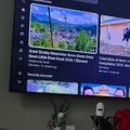 A TV screen displaying YouTube's recommended videos section, showing a video about Great Smoky Mountains with a scenic mountain town view, positioned above a shelf with decorative items including plants and flowers