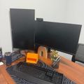 A desk workspace with two black computer monitors, a keyboard on a patterned mouse pad, and various desk accessories including an orange cube and rainbow decoration