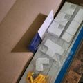 An open cardboard box containing white packaging materials, small white boxes, yellow plastic pieces, and instruction materials laid out on a wooden surface