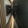 A flat-screen TV mounted on a wall bracket with visible cables, positioned against a wood-paneled wall in what appears to be a modern interior space.