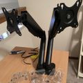 A black dual monitor arm stand assembled on a wooden desk with various screws and hardware pieces scattered around it