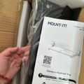A person's hand holding packaging materials and an instruction manual for a Mount-It clamp-on keyboard tray, with foam padding and cardboard box visible in the background