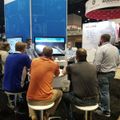 A group of people gathered around a presentation booth at a trade show or conference, with a presenter speaking to seated and standing attendees near display screens and company signage.