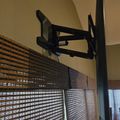 A black articulating TV wall mount attached to a pole near windows with horizontal blinds