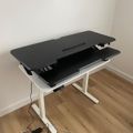 A height-adjustable standing desk with a black desktop surface and white base, positioned in a corner with wooden flooring and power cables visible underneath
