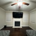A modern living room with gray walls, dark hardwood floors, white fireplace with mounted TV above, ceiling fan, and two gray tufted ottomans