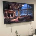 Wall-mounted TV displaying a cozy Christmas living room scene with fireplace, decorated tree, and snowy mountain view through large windows