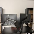 A dual monitor setup with articulating monitor arms on a dark desk, with shelving units and a white door visible in the background of what appears to be a home office space.