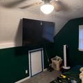 A room with green and white walls, gray carpet, and a ceiling fan with light fixture. A flat-screen TV is mounted on the upper wall, and various tools and equipment are visible on a green bed and floor, including a drill, measuring tape, and tool box.