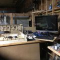 A cluttered workshop or basement workspace with wooden shelving, a computer monitor and keyboard on a desk, and various tools and equipment scattered around
