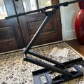 A black adjustable laptop stand or desk riser positioned on a patterned area rug in a home interior with wooden doors and decorative vase visible in the background
