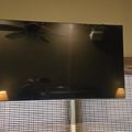 Large flat screen TV mounted on wall above bamboo window blinds with ceiling fan reflection visible on dark screen