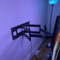 A room with purple and blue ambient lighting featuring a black adjustable TV wall mount, a tall floor lamp with blue illumination, and various cables on the floor near a baseboard.