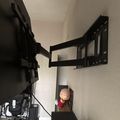 A black articulating TV wall mount bracket extended from a white wall in a corner, with various electronic equipment and cables visible below it, and a small figurine on a shelf in the background.