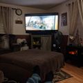 A cozy living room with a person's legs in jeans visible in the foreground, facing a large TV mounted on the wall showing a movie scene, with a brown couch, decorative curtains, and holiday decorations visible in the room.