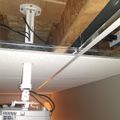 Ceiling-mounted projector installation with white mounting bracket, cables, and exposed ceiling framework visible above drop ceiling tiles
