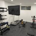 Home gym with treadmill, dumbbells on rack, exercise bike, wall-mounted TV, and gray carpeted floor in white-walled basement room