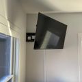 A flat-screen TV mounted on a wall bracket in a corner of a room with white walls, a window with blinds, and some items on a wooden surface below