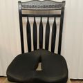 A black ergonomic seat cushion with a cut-out center placed on a wooden dining chair with vertical slats and decorative leaf pattern on the backrest