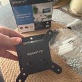 A person holding a black MOUNT IT! low-profile TV wall mount bracket with its packaging and hardware visible on a beige carpet. The box shows product images of the tilting mount installation.