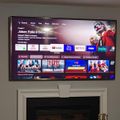 A wall-mounted TV displaying a streaming service interface with movie and show thumbnails, including titles like Stargate SG-1 and Criminal Minds, with various app icons visible at the top