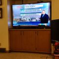 Television displaying a weather forecast with meteorologist presenting current conditions and upcoming weather information in a living room setting with wooden entertainment center