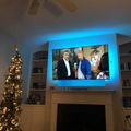 Living room with mounted TV showing two men, Christmas tree with lights, built-in shelving, and ceiling fan